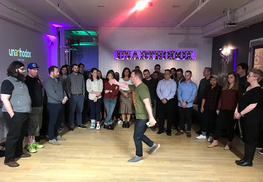 A group of people stands in a semi-circle at Unarthodox, while a man in the center gestures energetically